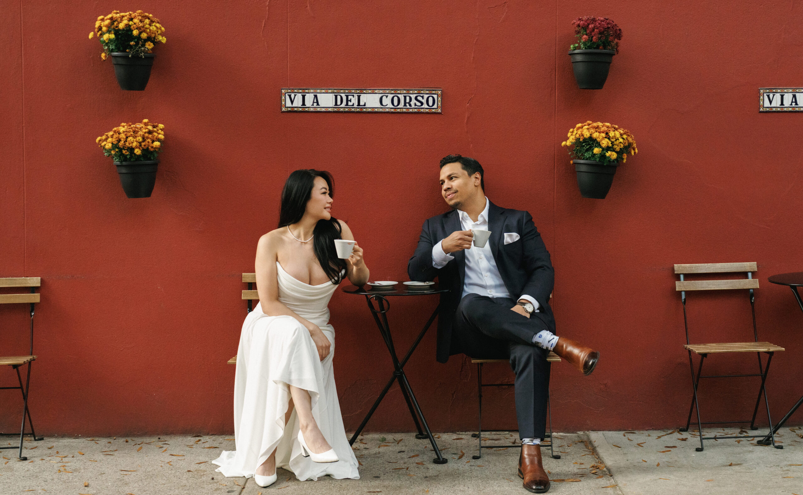 Bride to be and groom to be sitting at a bistro table drinking espresso against a red wall with hanging flower pots