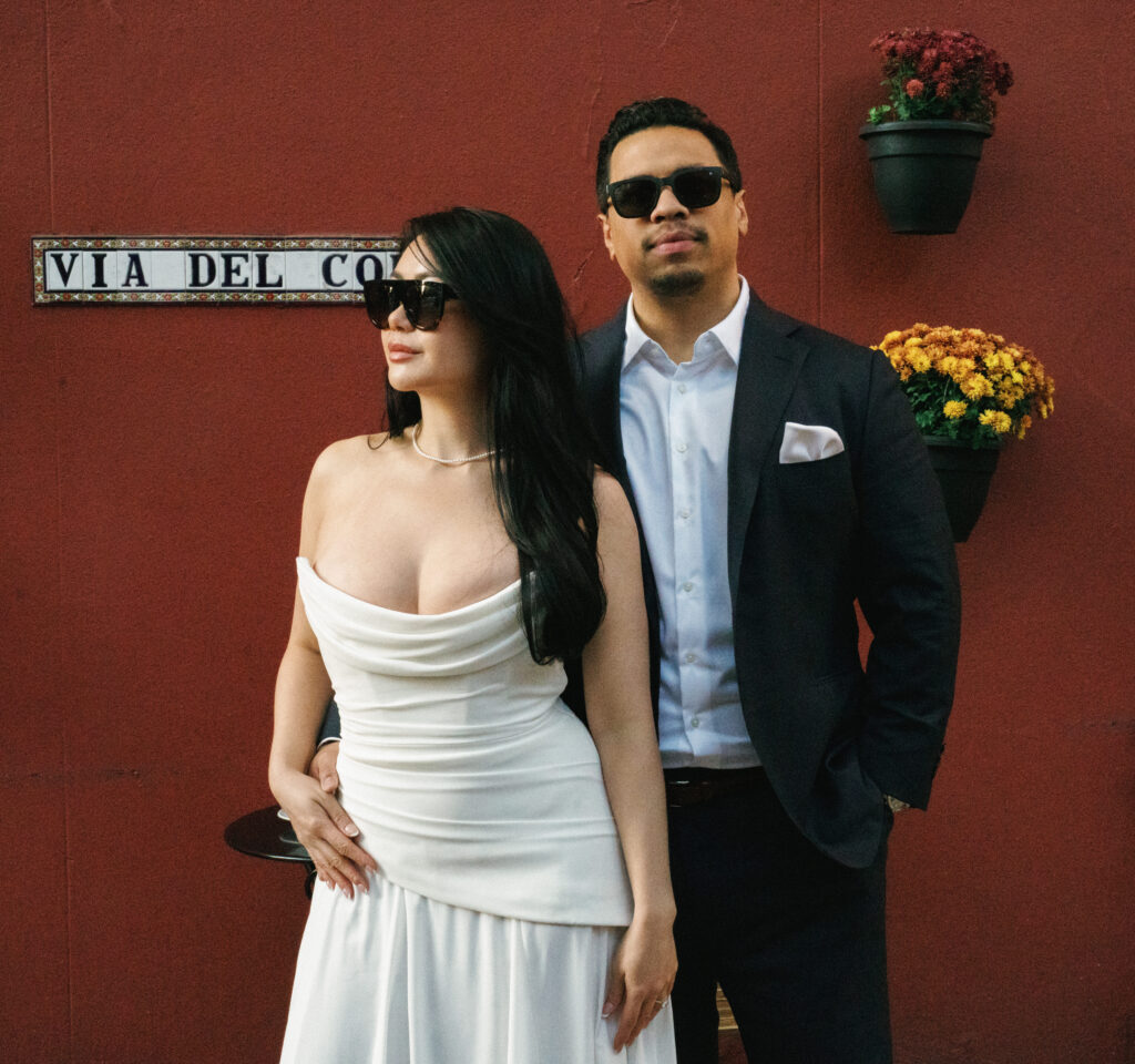 Groom to be standing behind his bride to be as they both look away in stylish black sunglasses