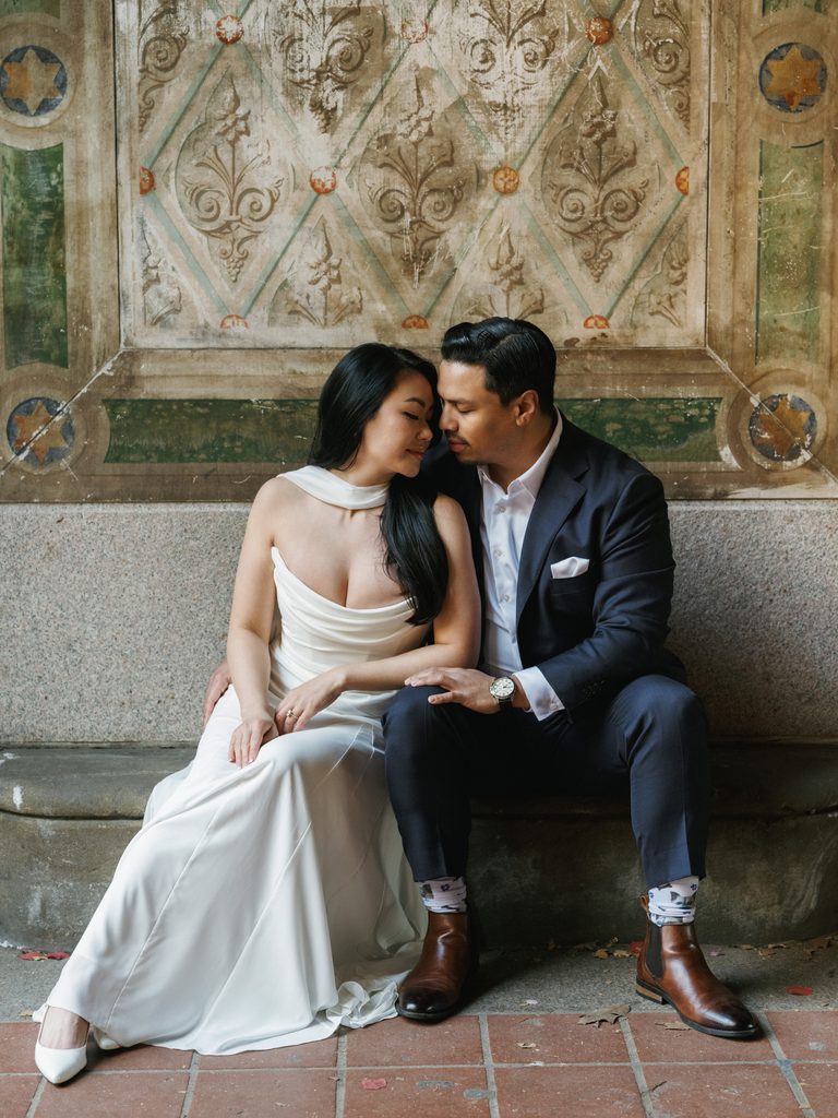 Bride-to-be in a white silk gown and groom-to-be in a navy suit sharing an intimate moment on a stone ledge.