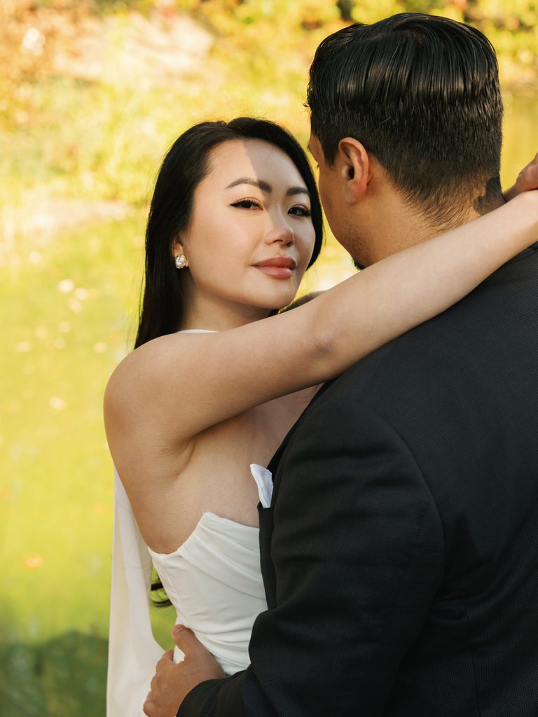 Bride to be looking over her fiance's shoulder during a romantic lakeside photo session in Central Park