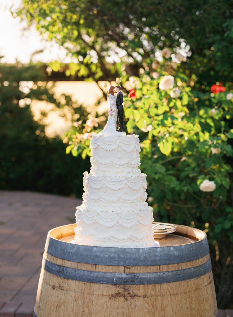 Elegant three tier vintage white wedding cake with custom bride and groom toppers standing on a rustic wooden wine barrel outdoor
