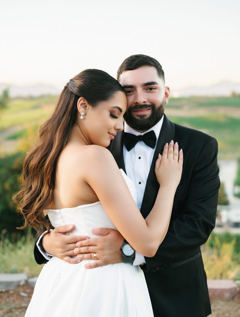 Intimate outdoor couple portrait of a groom in a classic black bow tie tuxedo hugging his bride showing her elegant half up half down wavy hairstyle and pearl drop earrings