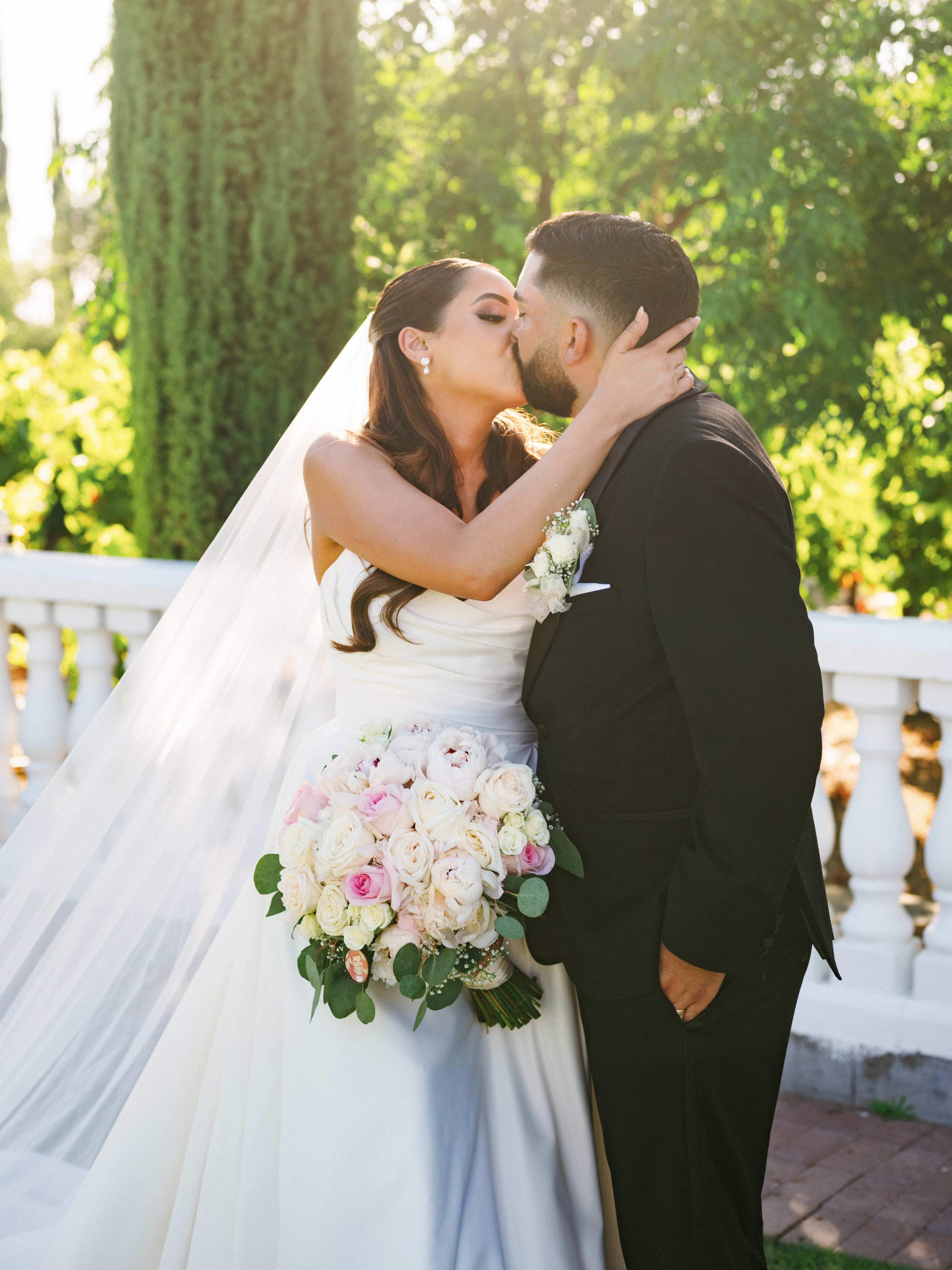 Cinematic romantic kiss of a luxury bride and groom near a white stone balustrade with a long flowing tulle veil in soft golden sunlight