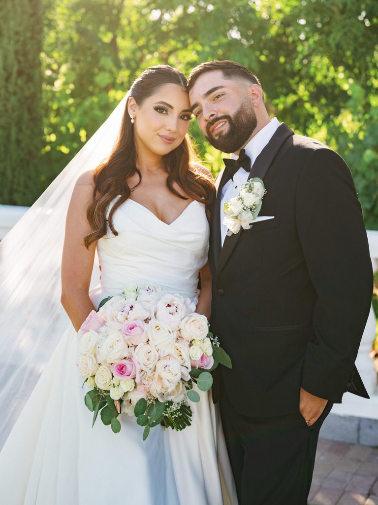 Sophisticated newlyweds posing outdoor with a lush white rose boutonniere and a stunning draped bodice bridal gown featuring a long tulle veil