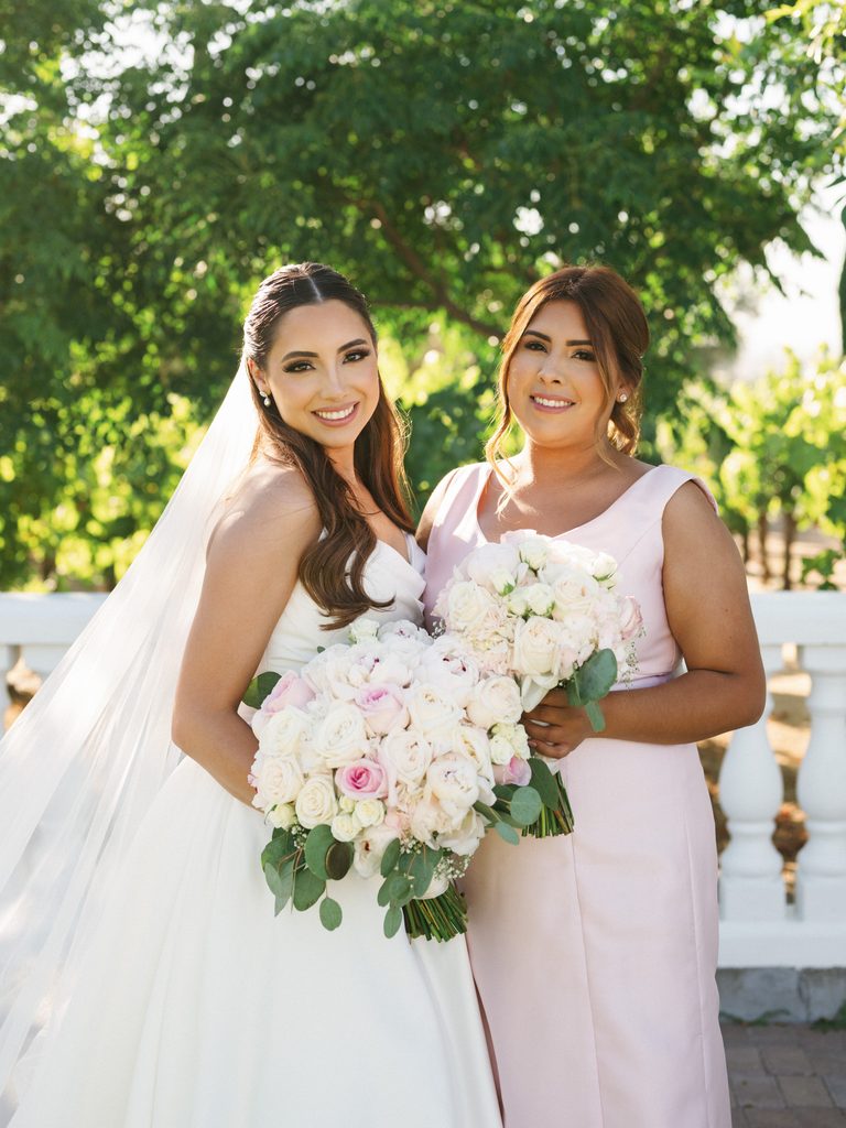 Gorgeous bride smiling with her bridesmaid in a chic blush pink sleeveless dress holding lush matching bouquets of white and pink garden roses