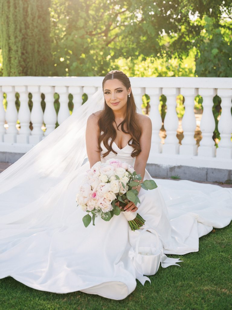 Stunning bride sitting on a green lawn with her voluminous white dress train and cathedral veil spread out holding a fresh garden flower bouquet