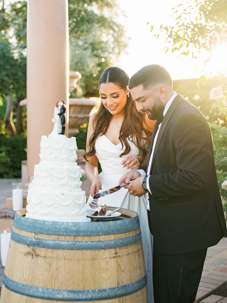Happy newlyweds cutting their luxury buttercream wedding cake on a vineyard reception terrace during a beautiful sunset golden hour