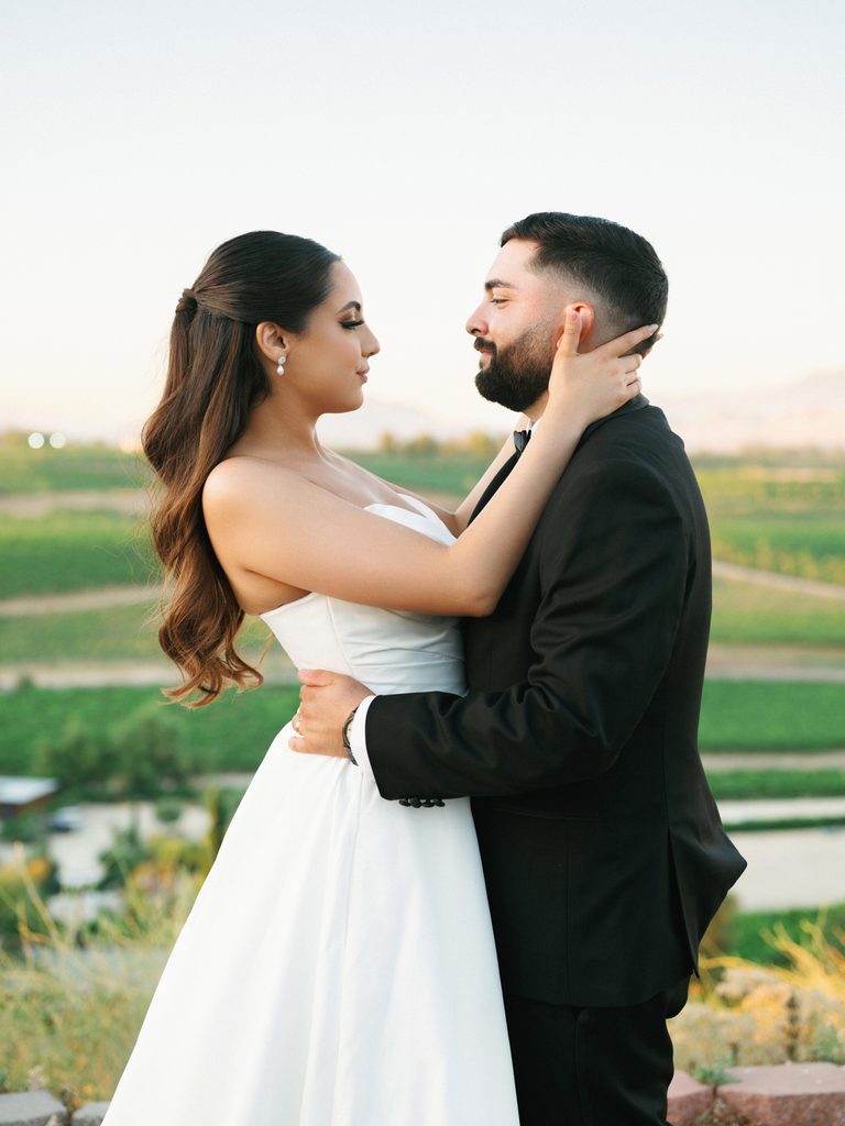 Elegant modern newlyweds posing outdoor while the bride gently touches her grooms face showing her classic half up half down wavy hairstyle