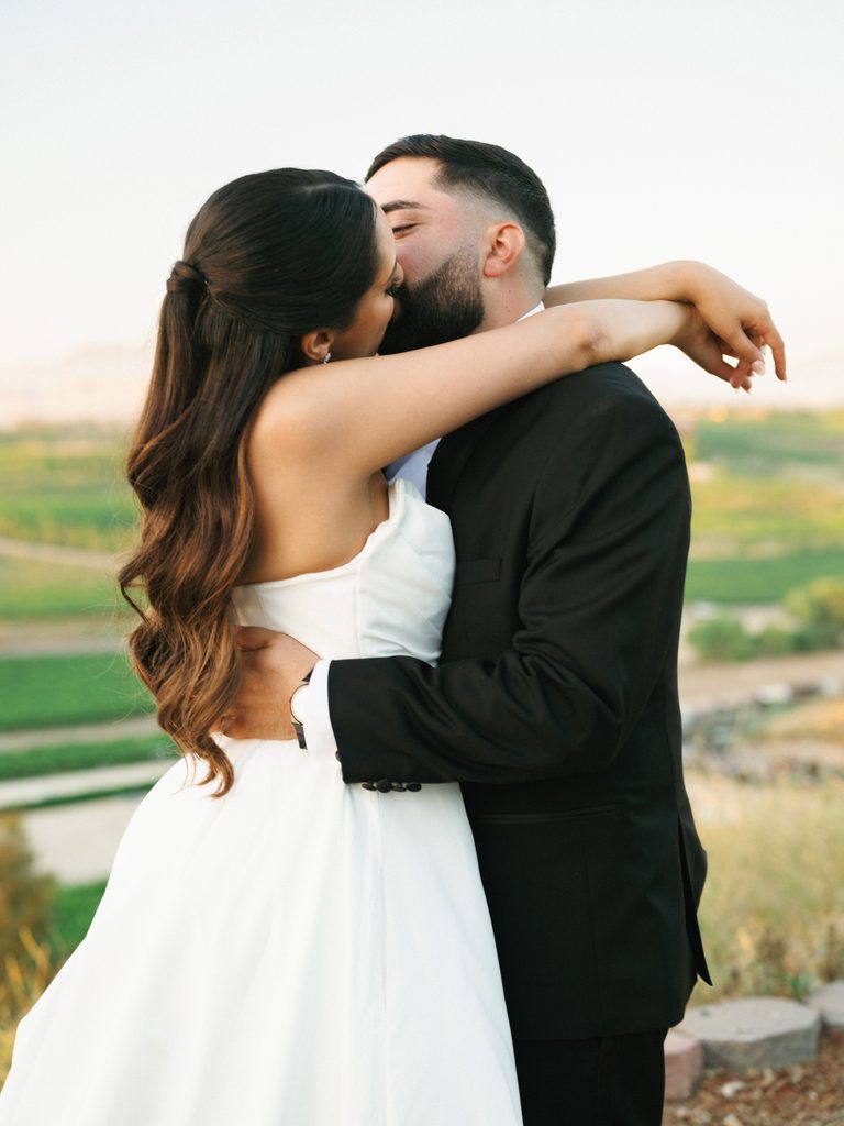 Cinematic wedding couple portrait of a groom hugging and kissing his bride against a breathtaking green vineyard landscape background