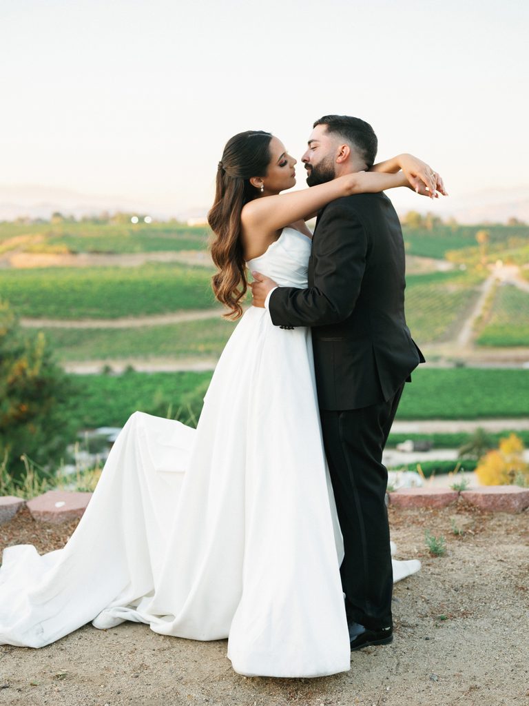 Breathtaking outdoor bridal couple portrait of a groom hugging his beautiful bride in a voluminous draped dress on a hill ridge overlooking scenic green valleys