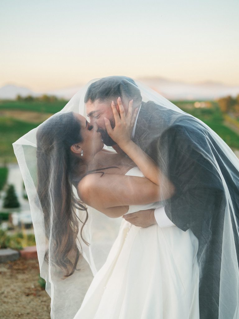 Intimate close up portrait of a bridal couple kissing under a sheer white veil while the bride gently touches her grooms face during a sunset photo session