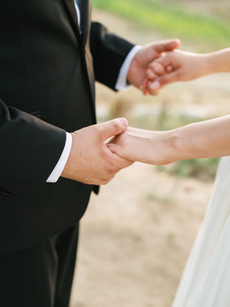 Close up portrait of a bride and groom holding hands outdoor with a sharp focus on emotional touch and texture of a black tuxedo sleeve