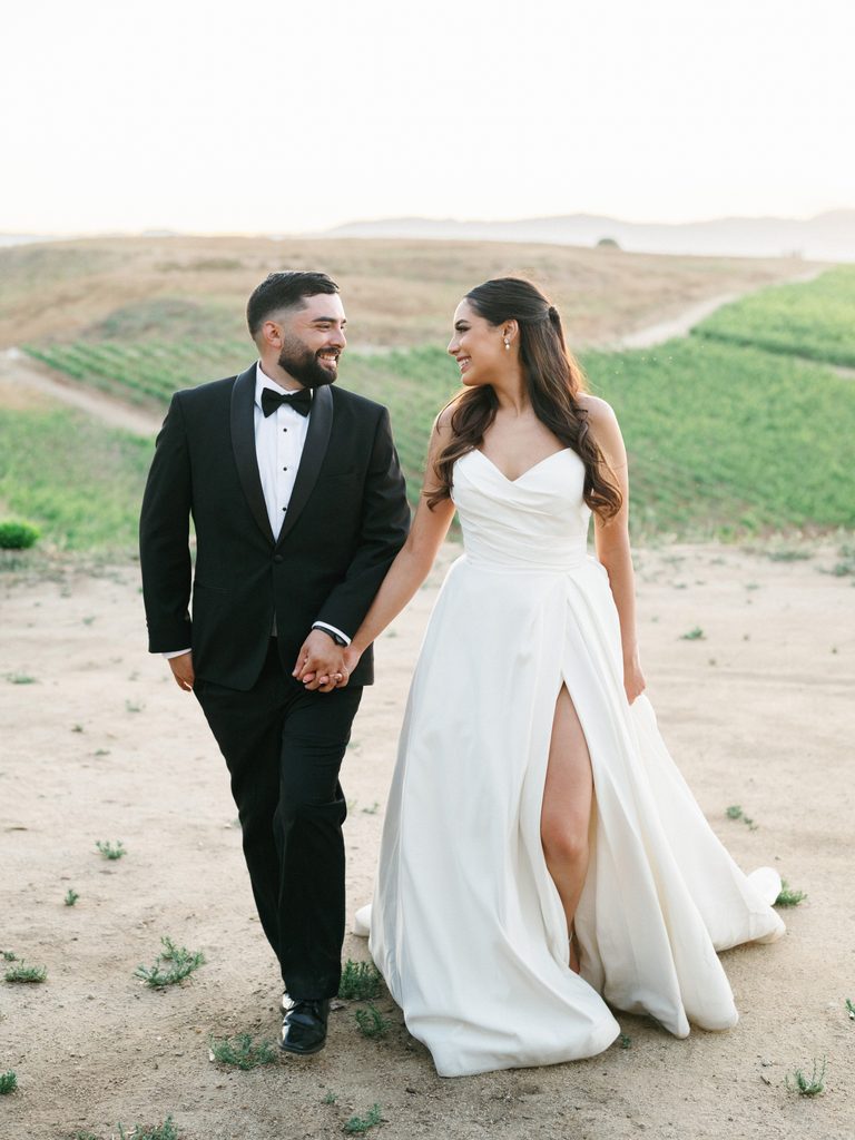 Joyful newlyweds walking hand in hand outdoor featuring a timeless black tuxedo and a chic draped bridal gown with a high leg slit