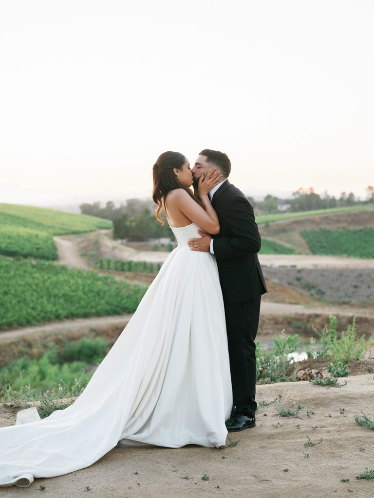 Breathtaking bridal couple portrait kissing on a scenic hill ridge overlooking beautiful green vineyard valleys featuring a long flowing white dress train