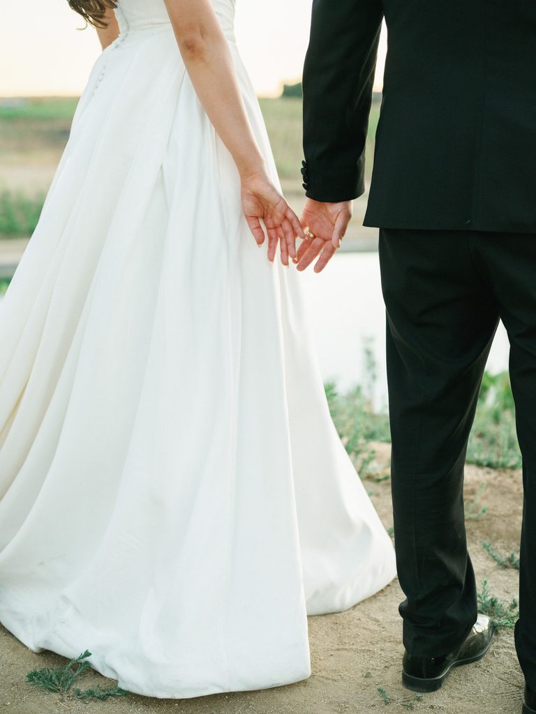 Close up of a bride and groom gently holding hands from behind showcasing a white silk ballgown skirt and sharp black tuxedo pants