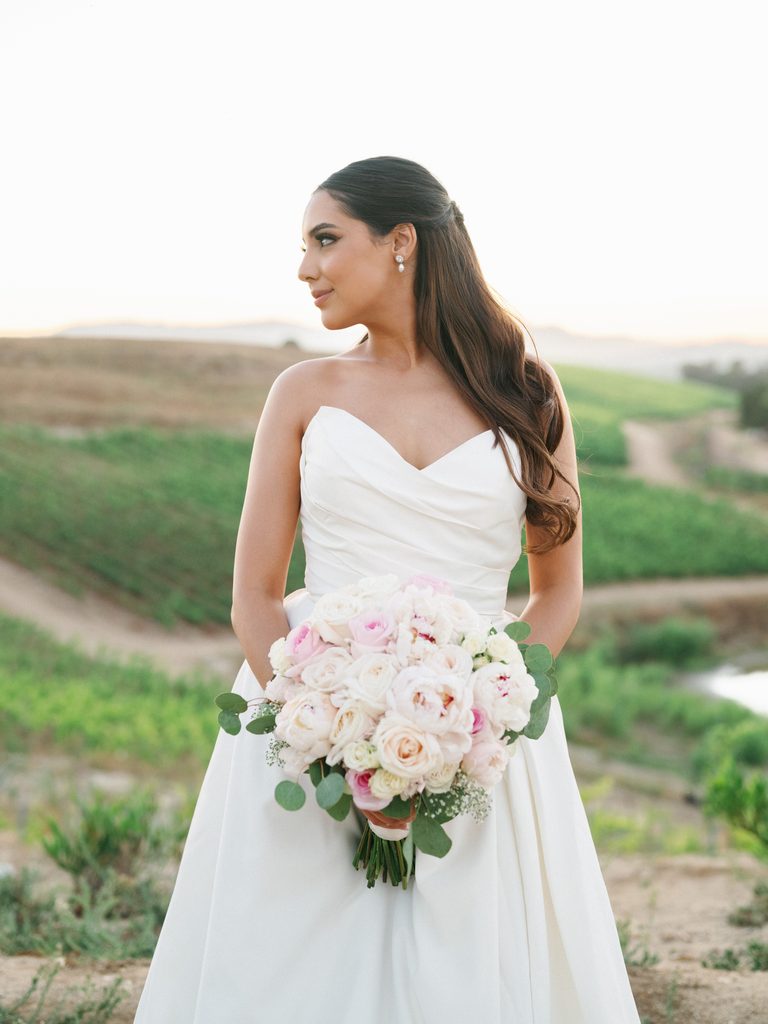 Elegant bride holding a lush white and pink rose bouquet looking away to the side showing her classic wavy half up half down hairstyle