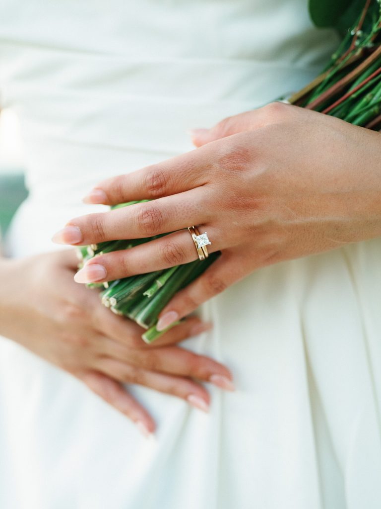 Close up of a brides hand wearing a luxury princess cut diamond engagement ring with a matching gold wedding band holding flower stems