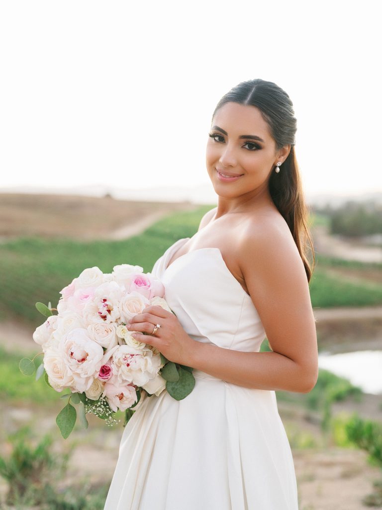 Stunning bride in a strapless silk dress posing outdoor on a hill holding a lush round bouquet of white garden roses and pink peonies