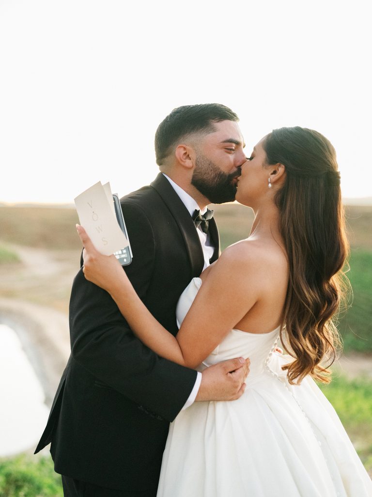 Romantic newlyweds sharing a sweet kiss outdoor while the groom holds an elegant minimal wedding vow book during a scenic vineyard session