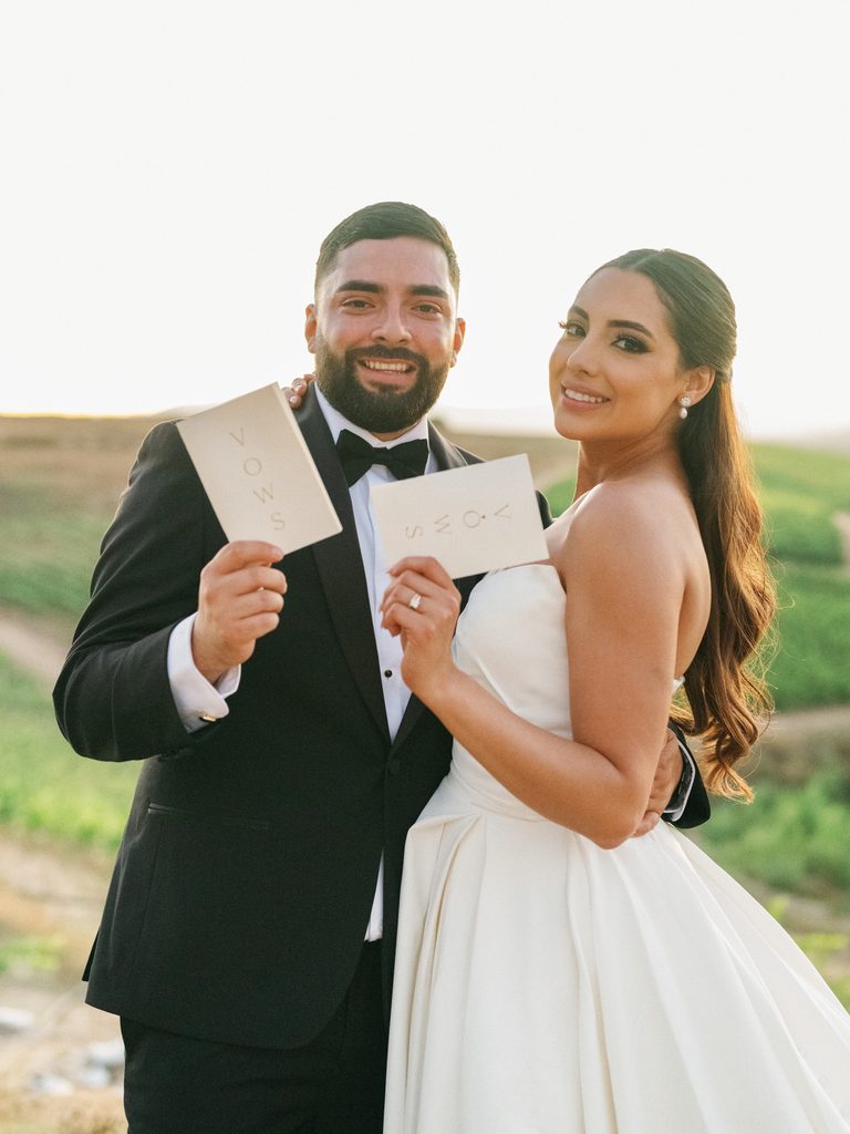 Happy bride and groom smiling and holding up matching chic white wedding vow booklets posing on a beautiful sunlit hill