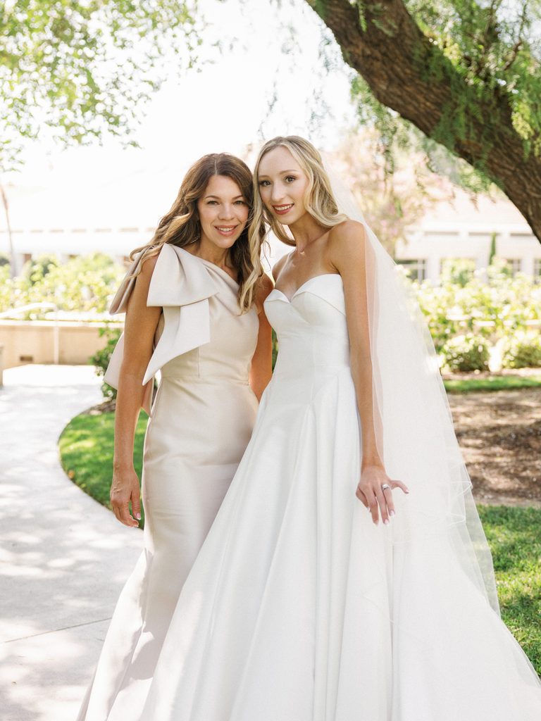 Beautiful blonde bride posing with her mother wearing a chic champagne color column dress with an oversized shoulder bow detail