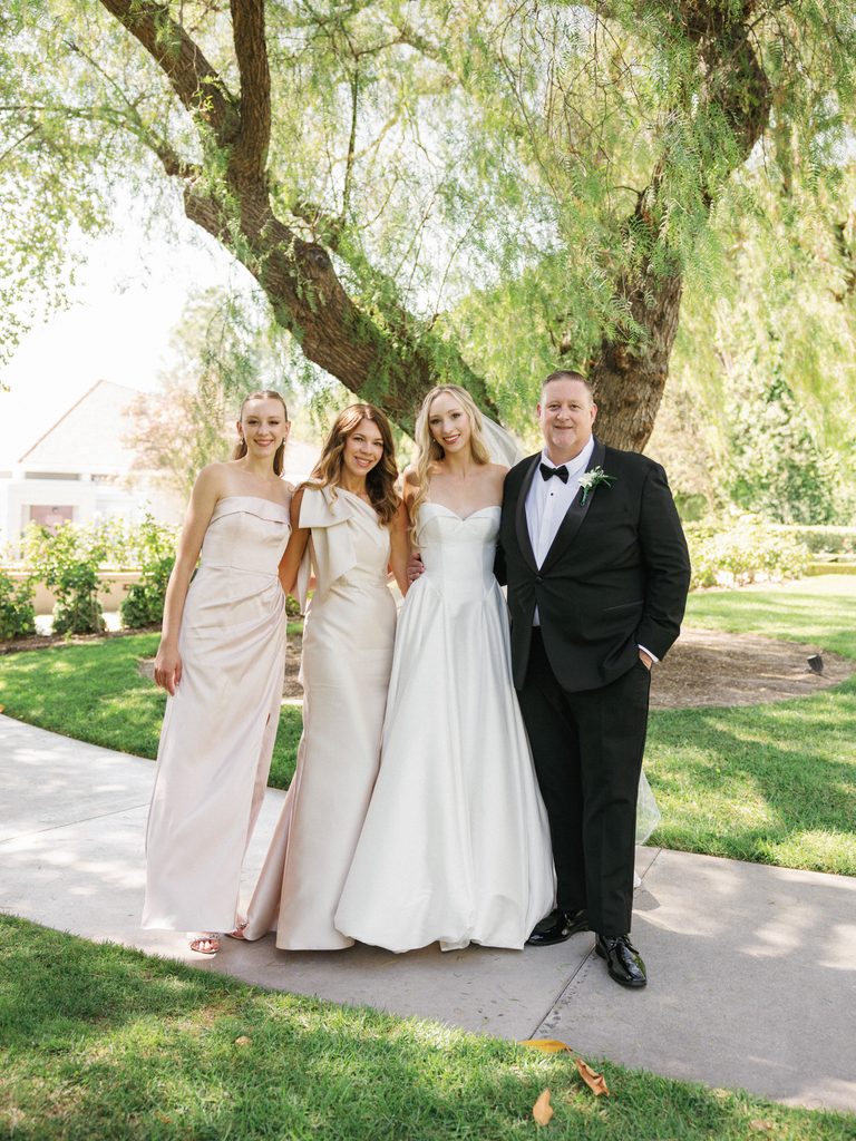 Elegant family wedding portrait outdoor on a paved stone pathway under a large tree featuring stylish bridesmaids and parents