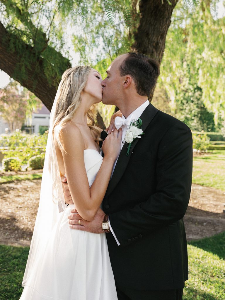 Romantic newlyweds sharing a passionate kiss outdoor featuring a classic black tuxedo with a white bow tie and elegant bridal makeup