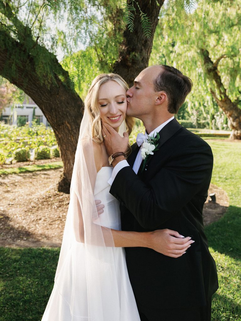 Sophisticated groom gently kissing his smiling brides cheek during an intimate wedding portrait session under large green trees