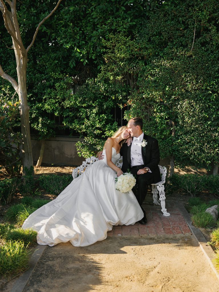 Chic newlyweds sitting together on a white ornate wrought iron garden bench surrounded by lush green trees and warm sunlight
