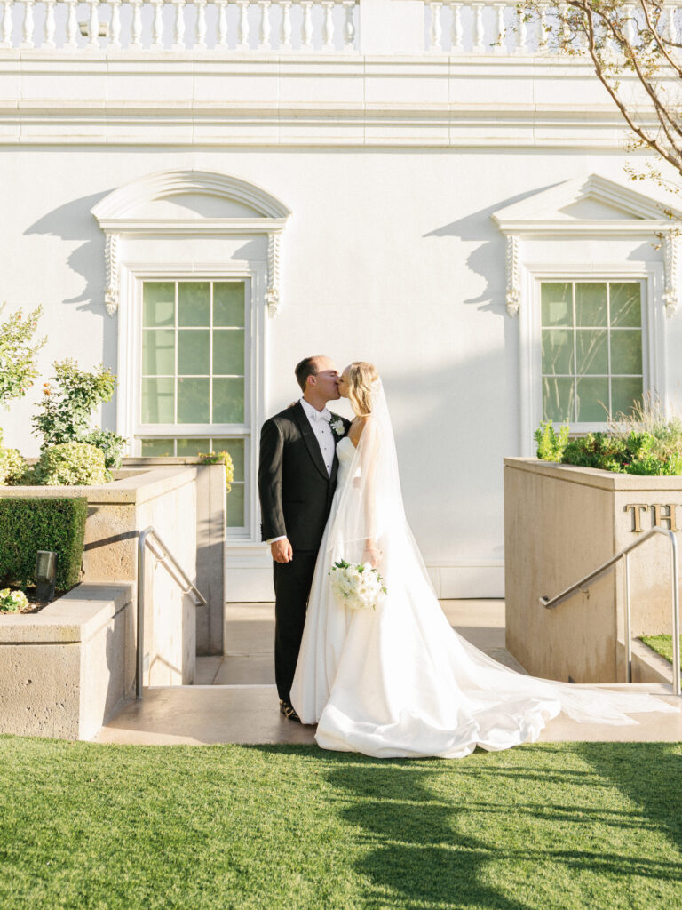 Cinematic wedding day couple kiss in front of a grand white estate building with elegant arch windows and manicured green lawn