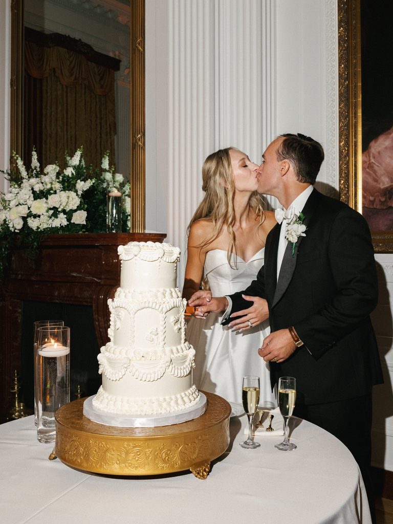 Chic newlyweds sharing a romantic kiss while cutting a three tier vintage white wedding cake on an ornate gold stand
