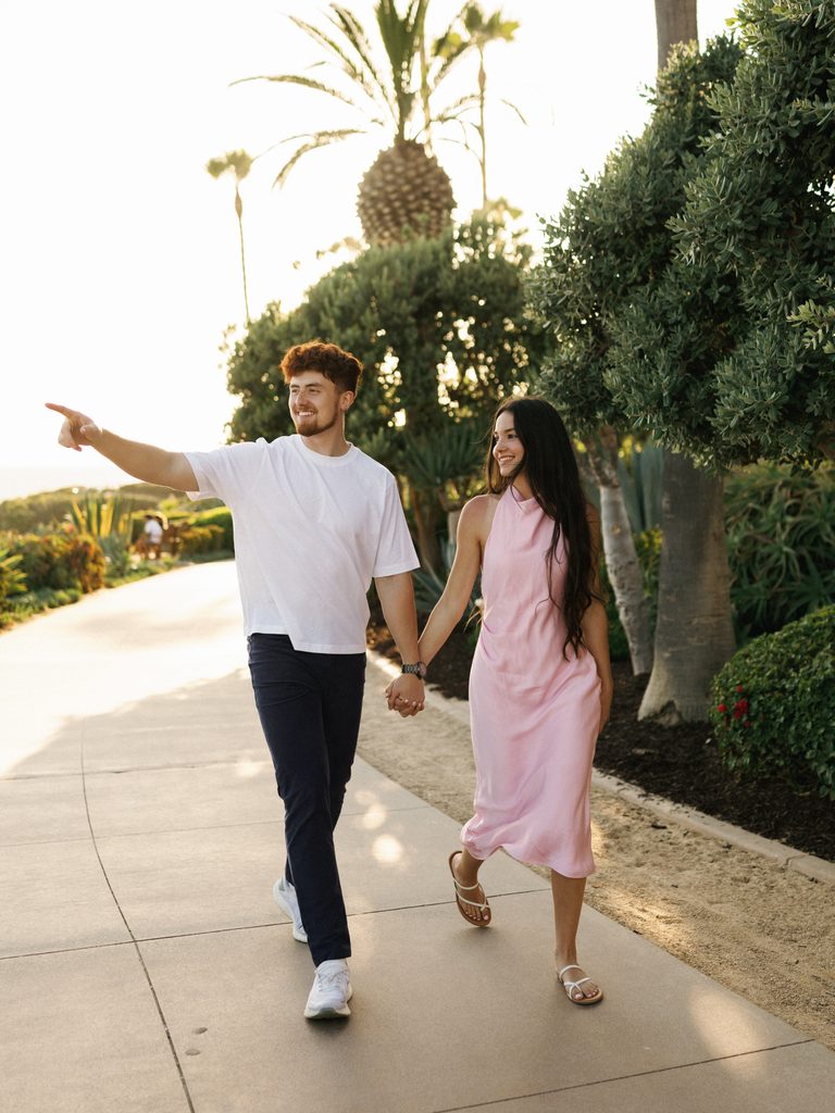 Man in a white t-shirt and bride-to-be in a pink halter midi dress walking on a path, showcasing casual resort-style engagement photo outfits.