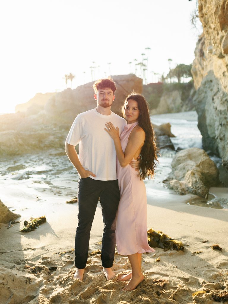 Barefoot couple standing on the sand by rocky cliffs during a sunset coastal photoshoot.