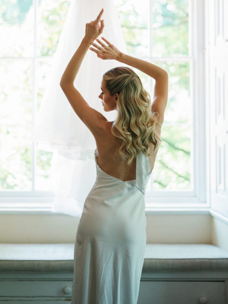 Graceful bride standing by a bright window shows off the open back details of her reception dress
