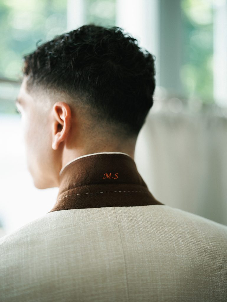 Groom from behind reveals custom embroidered initials under the collar of his tailored beige wedding suit