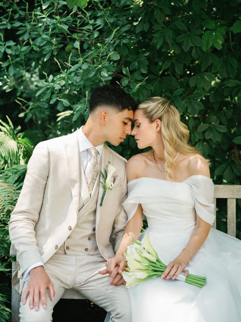 Newlyweds share an intimate moment on a garden bench leaning in close with closed eyes