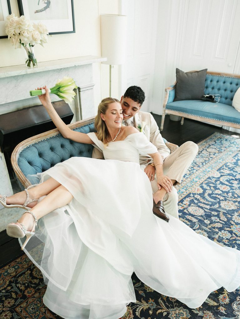 Playful bride and groom laughing together while relaxing on a blue tufted sofa inside a luxury estate