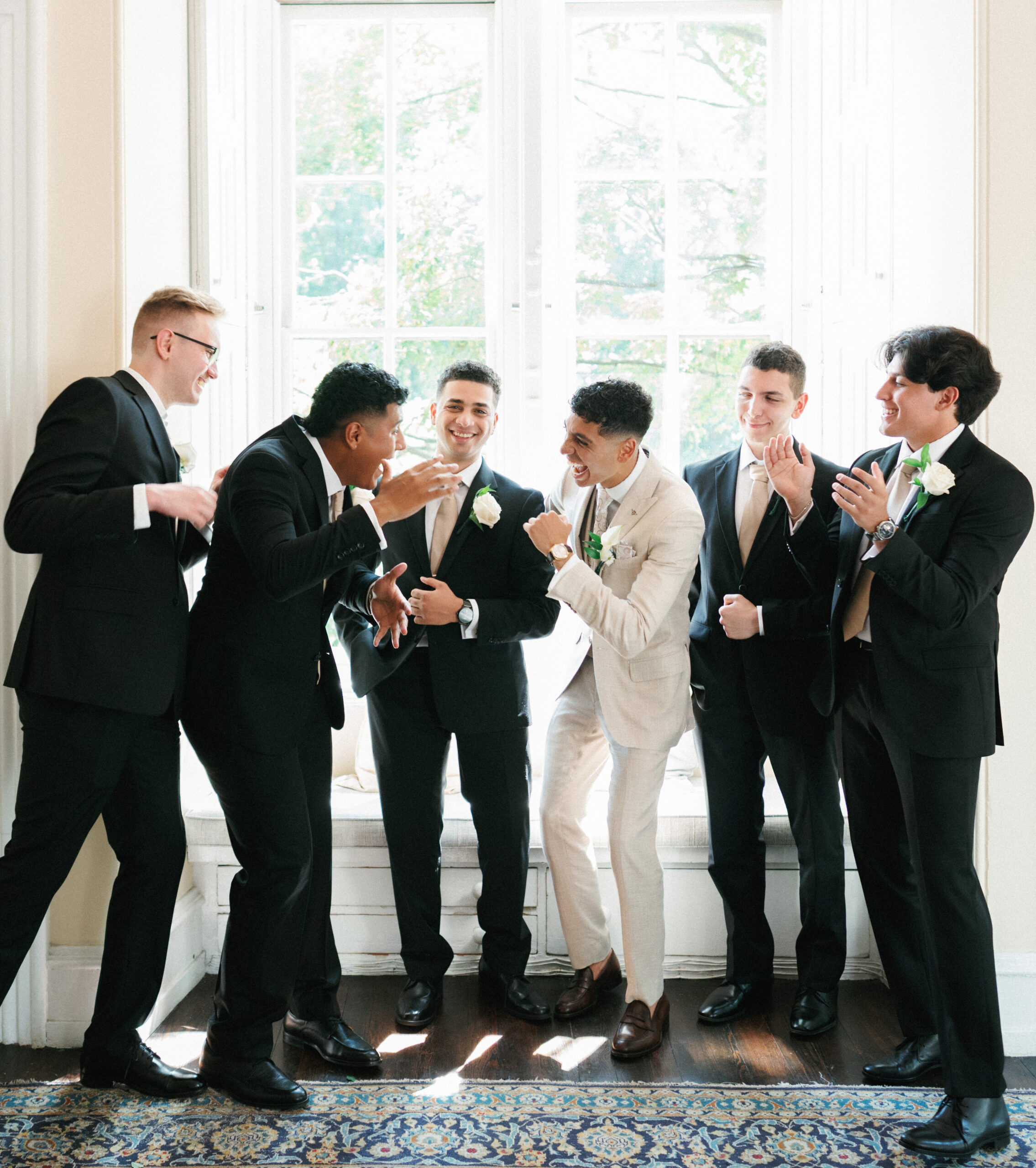 Groom and groomsmen share a genuine laugh while getting ready by a bright window inside a luxury estate
