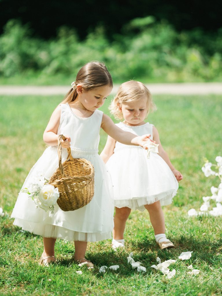 Two sweet flower girls scatter white flower petals on the grass at an upscale outdoor wedding venue