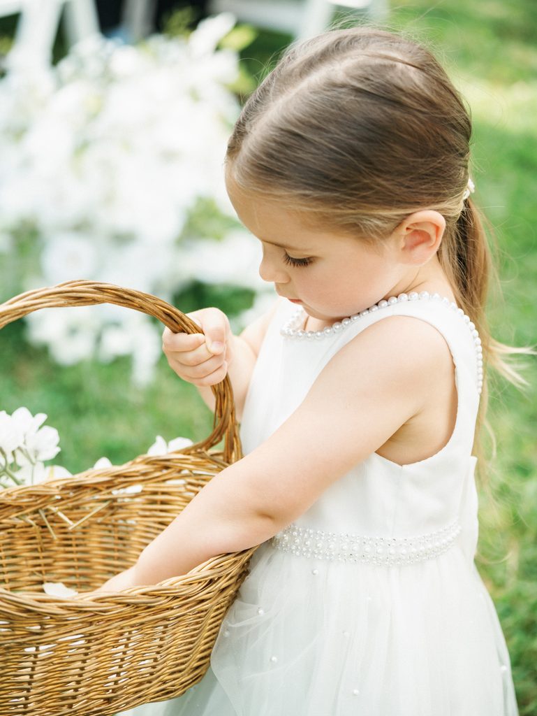 Adorable flower girl in a white pearl detailed dress looks into her woven basket during an outdoor ceremony
