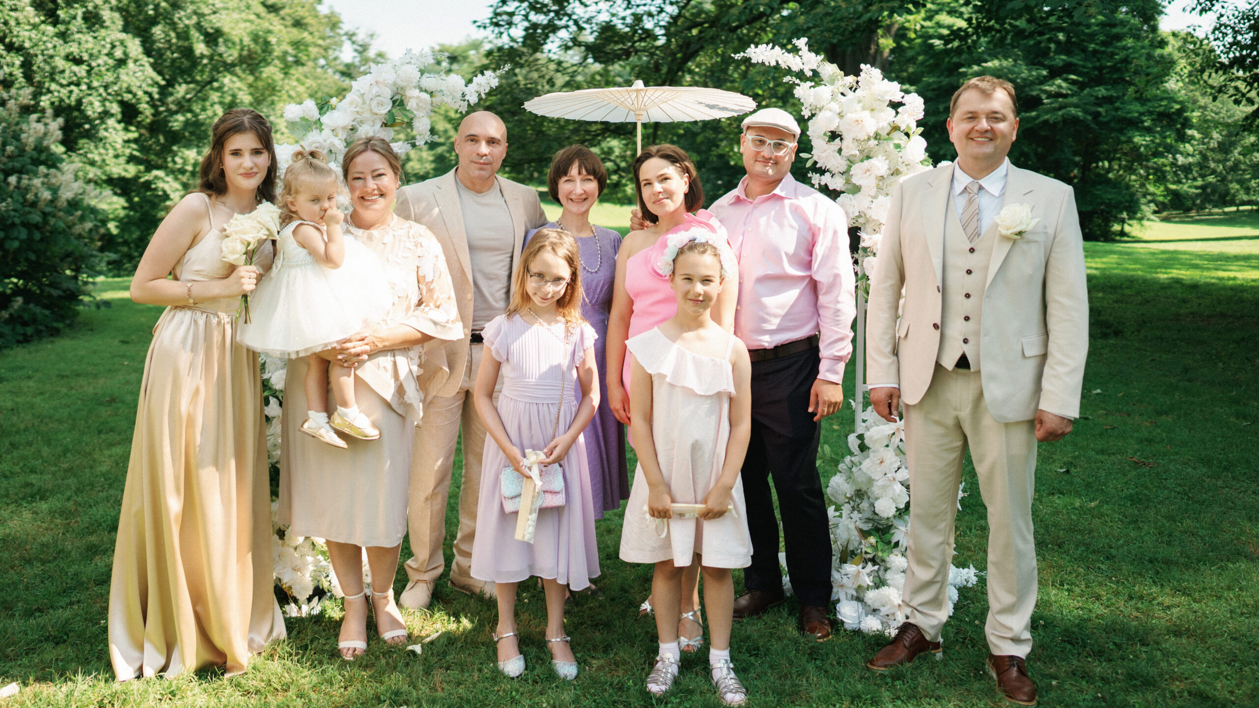 Smiling family members and wedding guests pose together in front of a white floral arch during an outdoor garden ceremony