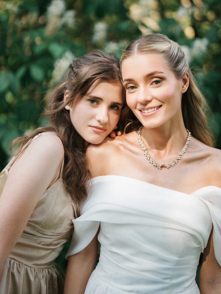 Beautiful bride smiles next to her maid of honor during an outdoor portrait session in a lush green garden