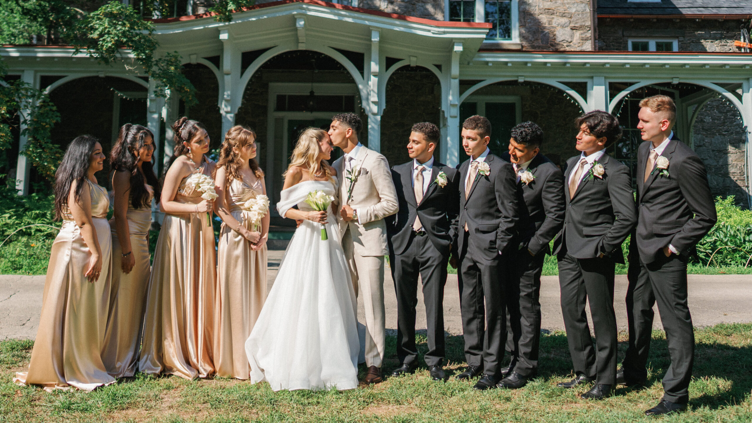 Bridal party stands together on a lawn smiling at the newlyweds kissing in front of a luxury historic estate