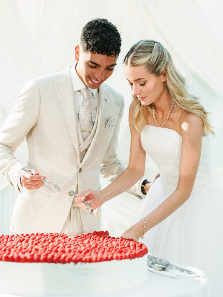 Newlyweds look down smiling while cutting a stunning cake topped with fresh red berries