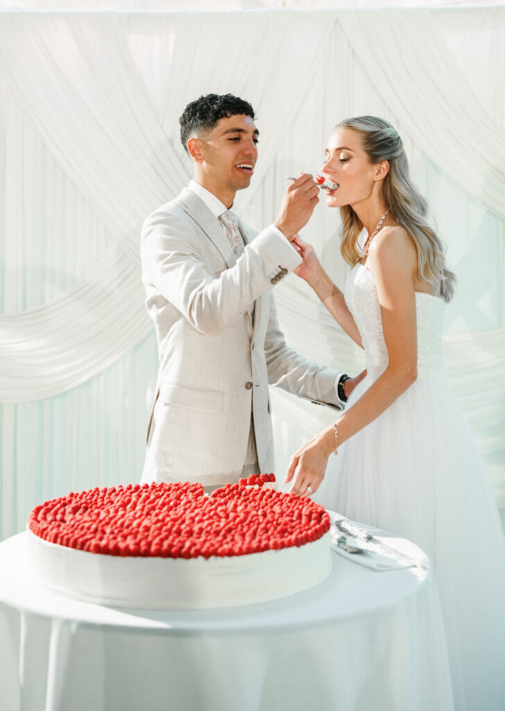 Groom feeds the bride a bite of their modern raspberry wedding cake during a luxury reception
