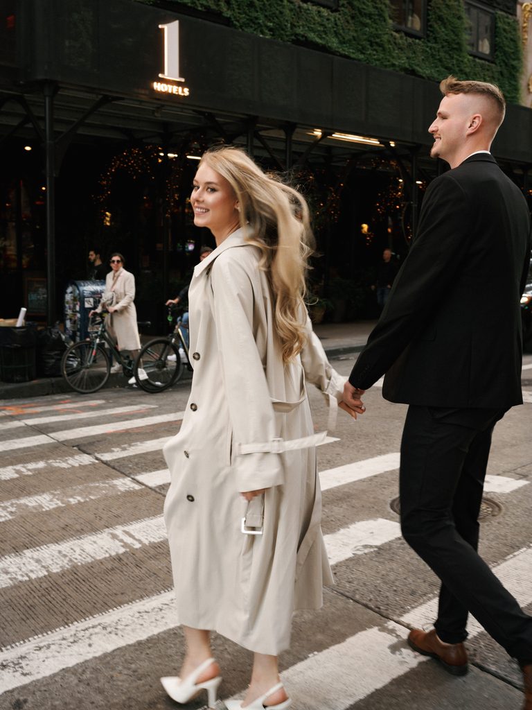 Bride-to-be in a beige trench coat looking back at the camera while holding hands with her fiancé, inspiring city-chic engagement photo outfits.
