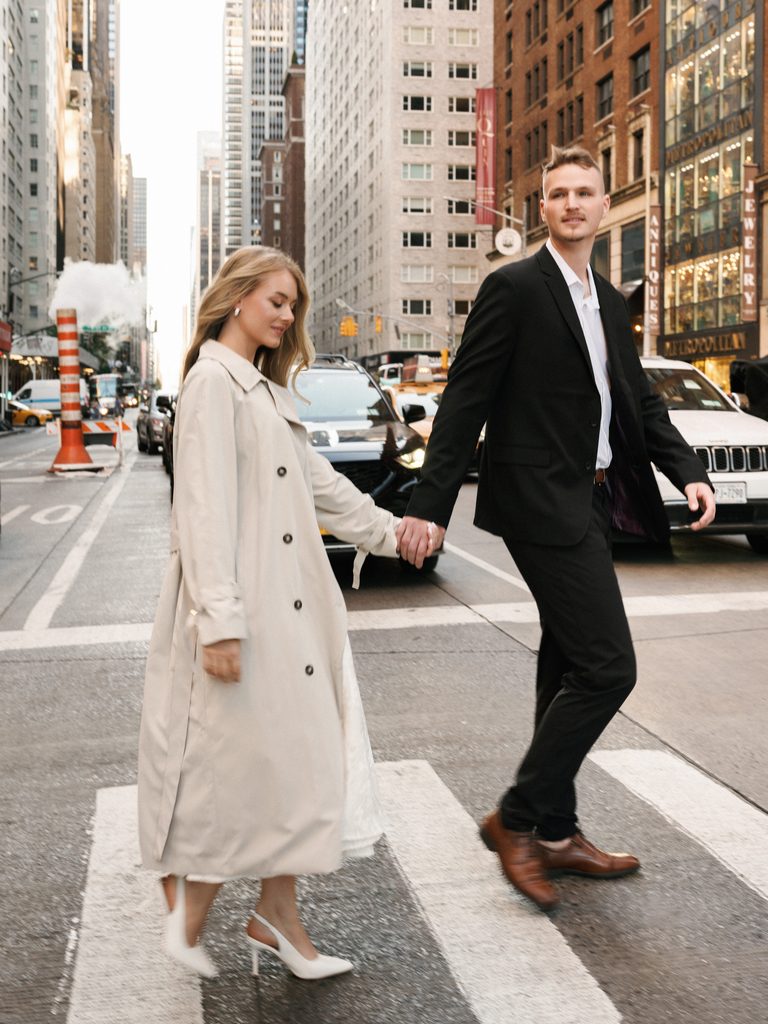 Man in a black suit and a bride-to-be in a long cream trench coat walking on a crosswalk surrounded by city skyscrapers.