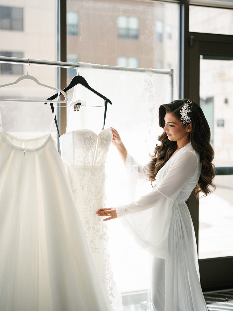 Elegant future bride in a white silk morning robe looking at her luxury couture wedding gown on a clothing rack
