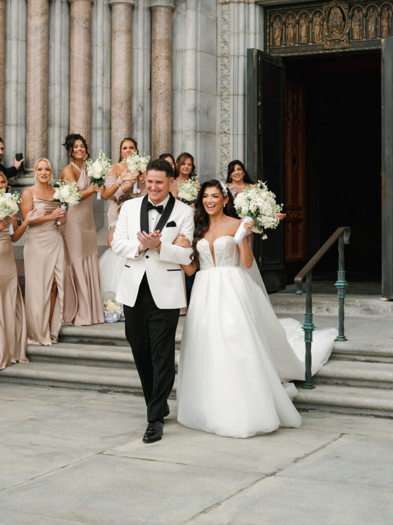 Excited newlyweds walking down cathedral stairs with bridesmaids cheering during a high end event
