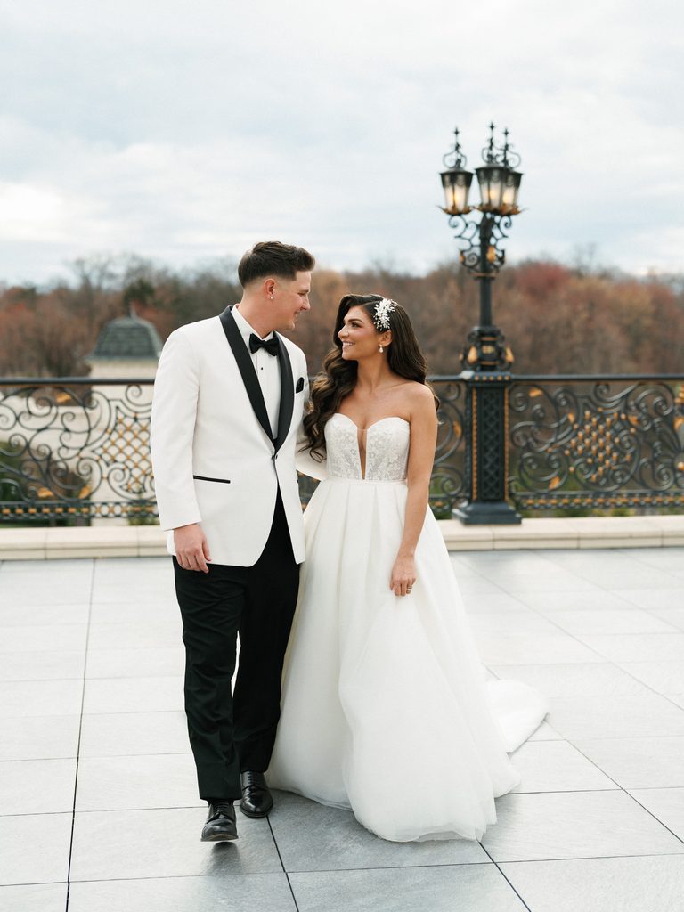 Editorial bridal couple smiling at each other during an outdoor stroll featuring an upscale white tuxedo jacket and flowing a line wedding gown